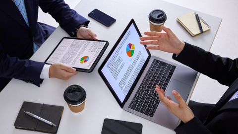 Two professionals seated in front of their laptops that are opened to a screen displaying a pie chart and text.