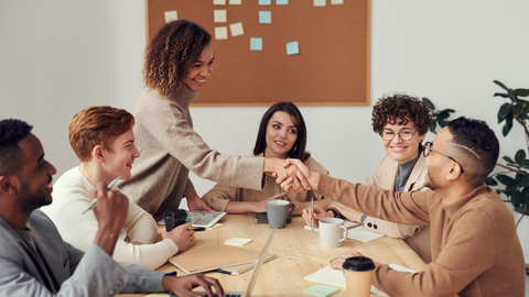 Woman standing at a table filled with people reaches out and shakes hand of man who is sitting down.