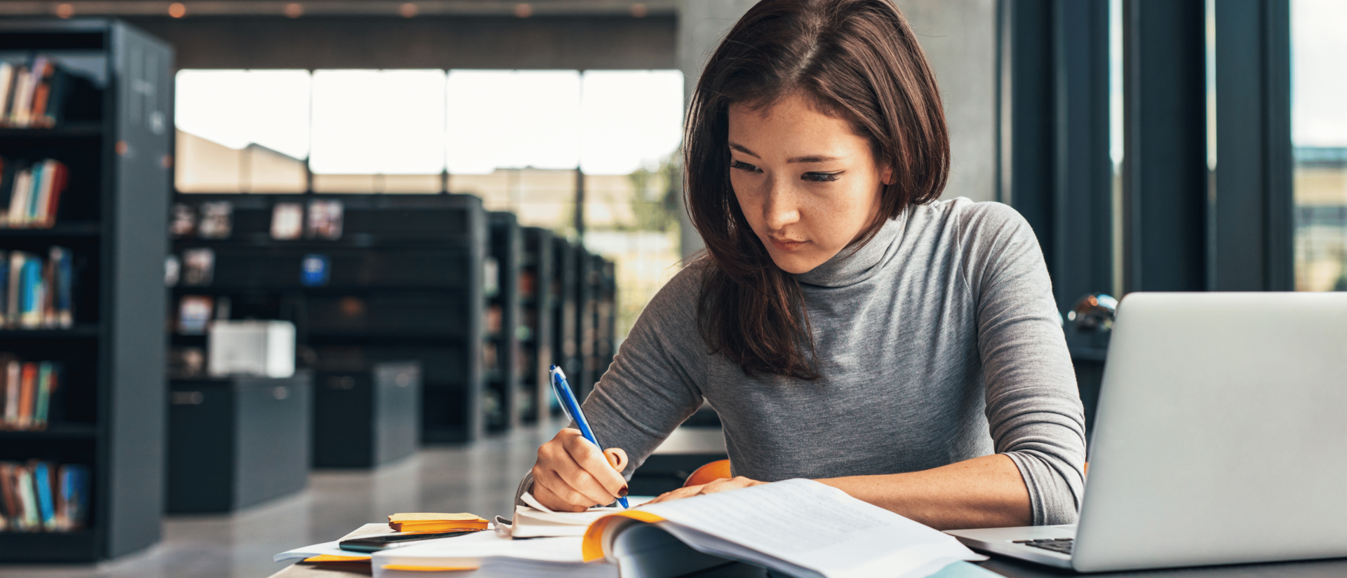 Woman in library with open book on table as she writes notes.